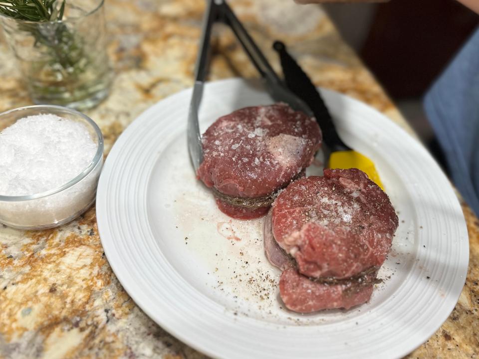 Two raw filet mignon steaks sprinkled with salt and pepper on a white plate, with tongs around one of the steaks. A bowl of salt and a glass with rosemary sprigs sit next to the plate