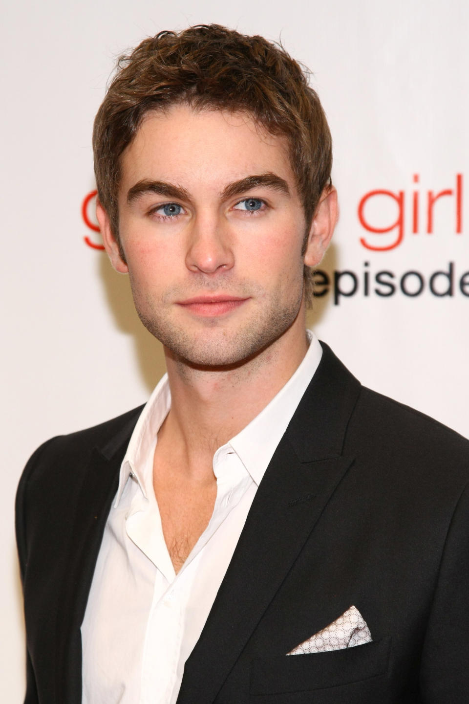 Chace Crawford at a red carpet event wearing a black suit jacket over an open white dress shirt