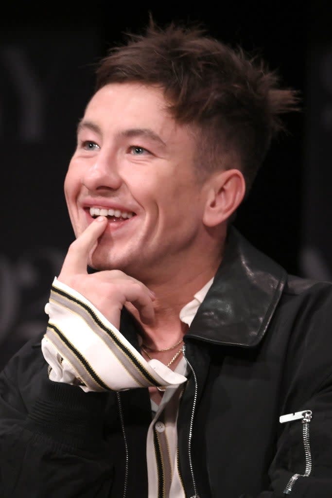 Barry Sloane wearing a black leather jacket with white stripes, smiling with hand on chin at an event