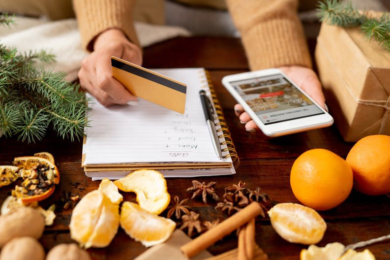 A woman writing a gift list while holding a credit card and phone with oranges and spices on the table in front of her.