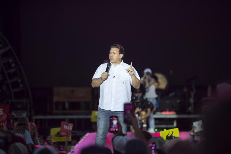 Ron DeSantis stands onstage lit by spotlight before a crowd and holds a microphone with his right hand.