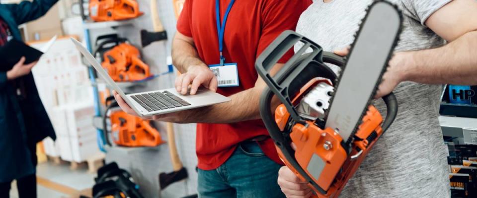 Salesman in red shirt and baseball cap is showing bearded client new chainsaw explaining details on laptop in power tools store.