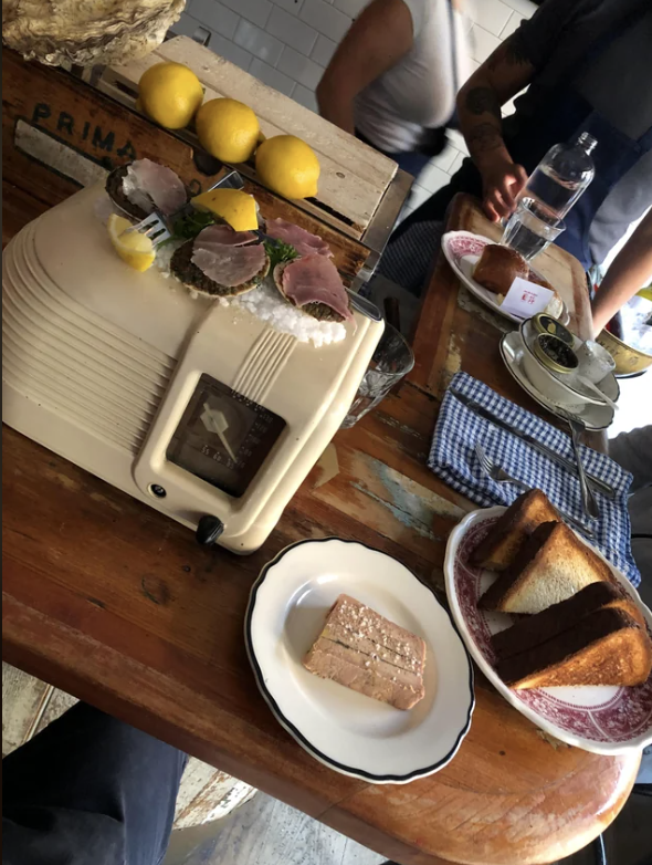 A vintage radio sits on a wooden table. On the table are lemons, sliced bread, two plates, and a wrapped butter pat. In the background, two people are dining