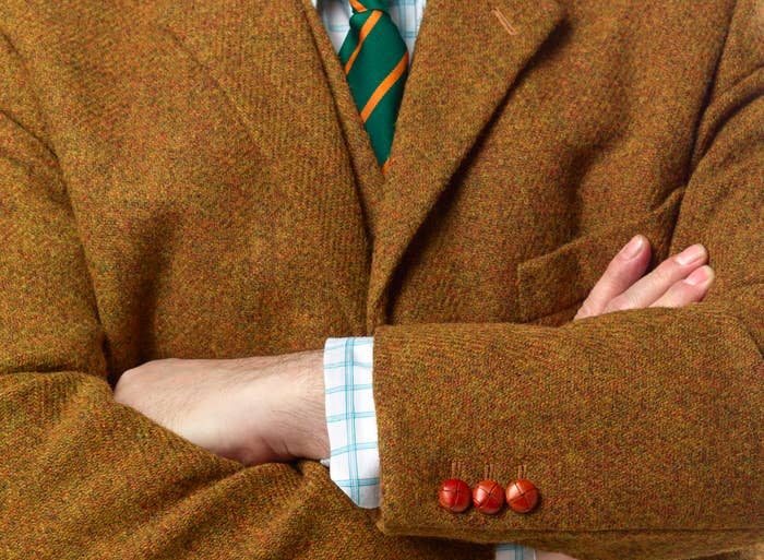 Person with arms crossed, wearing a brown tweed jacket, a green-striped tie, and a plaid shirt. Face not shown