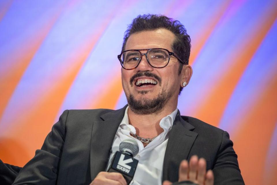 john leguizamo wearing a black suit jacket and white shirt, wearing glasses, speaking into a microphone