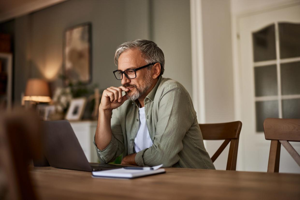 A focused mature male freelancer sitting at the wooden table at home and reading online projects on a laptop.