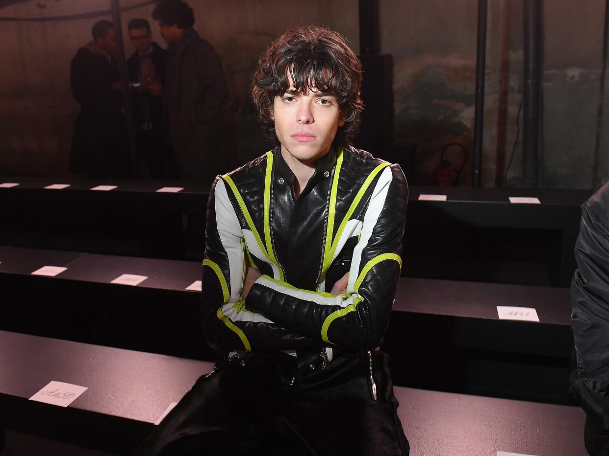 Luca Fersko sitting down on a bench before a fashion show, with his arms crossed.