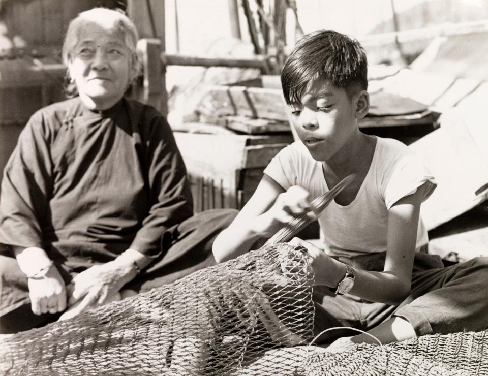 A boy repairs fishing nets in Aberdeen Harbor as an elderly person behind them looks into the distance.