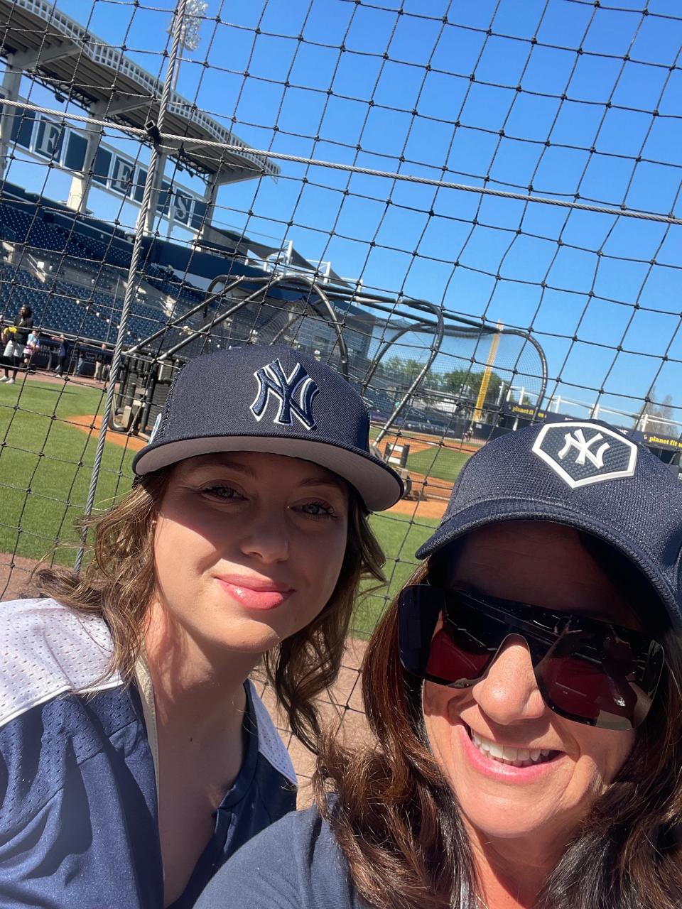 Two women in Yankee hats stand in front of a baseball field for a picture.