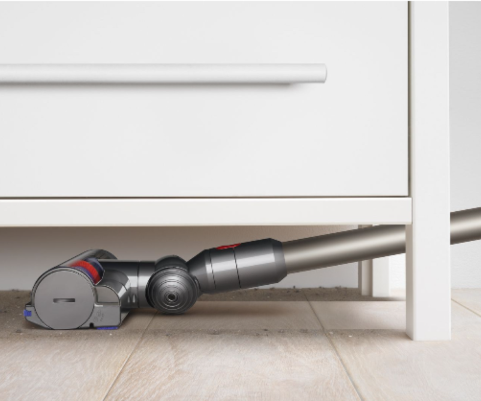 A close up of a grey Dyson vaccum head cleaning the floor under a white cabinet.