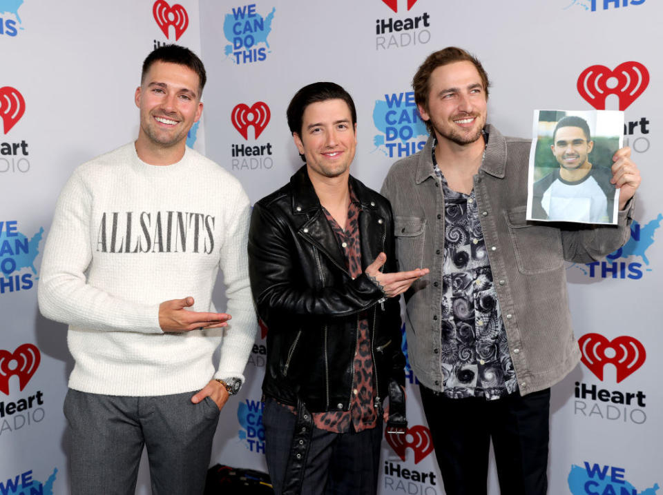 Three smiling members, with one holding a photo of the fourth, dressed casually in long-sleeved sweaters, jackets, vests, or shirts