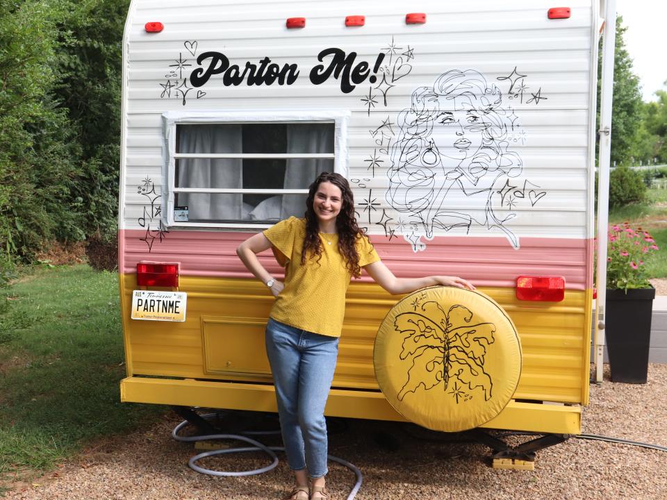 Reporter Talia Lakritz, wearing a yellow shirt and jeans, stands in front of a Dolly Parton-themed RV that says "Parton Me!" on the back.
