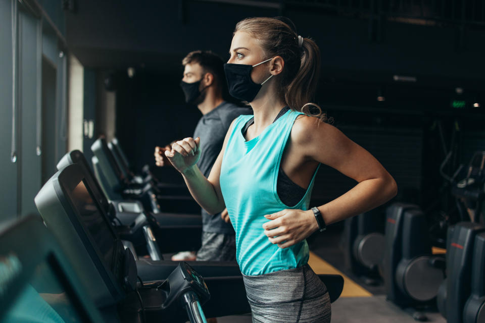 Young fit woman and man running on treadmill in modern fitness gym. They keeping distance and wearing protective face masks. Coronavirus world pandemic and sport theme.