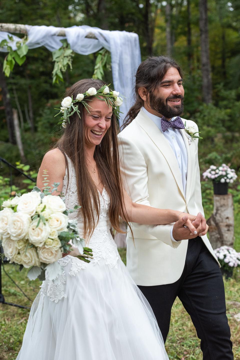 Anthony and Kelly Anne Ferraro at their wedding. The bride wears a white dress and a flower crown and carries a bouquet. The groom wears a white tux and black pants with a purple bowtie.