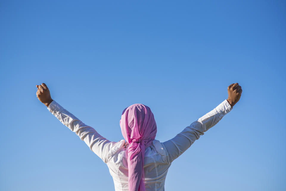 Person raising their arms in a triumphant gesture against a clear sky, wearing a headscarf and a long-sleeve shirt, viewed from behind