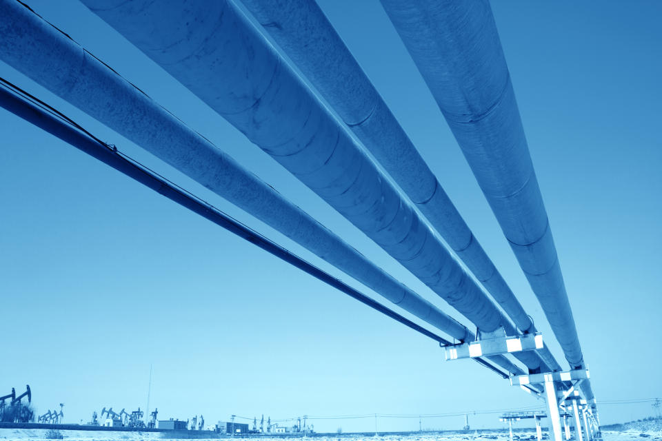 Several pipelines overhead with a blue background and oil pumpjacks in the foreground.