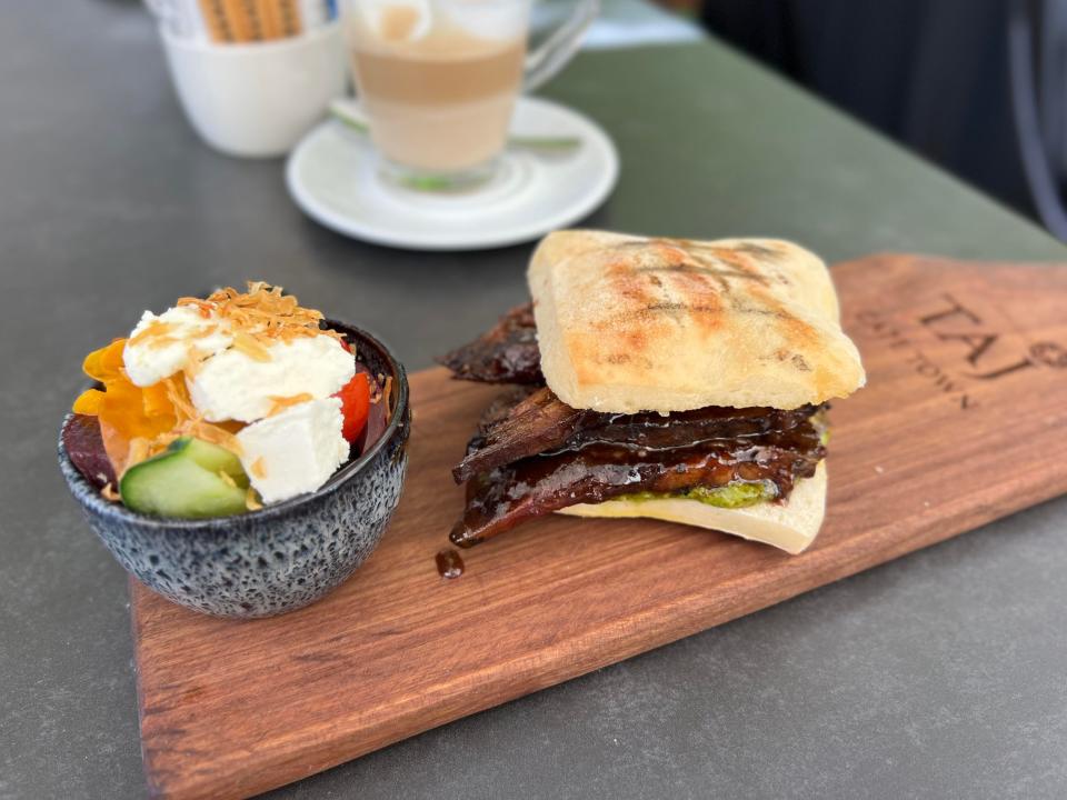 A paddle engraved with "Taj Cape Town" holding a brisket sandwich and a gray cup of a salad with cheese and cucumbers in it