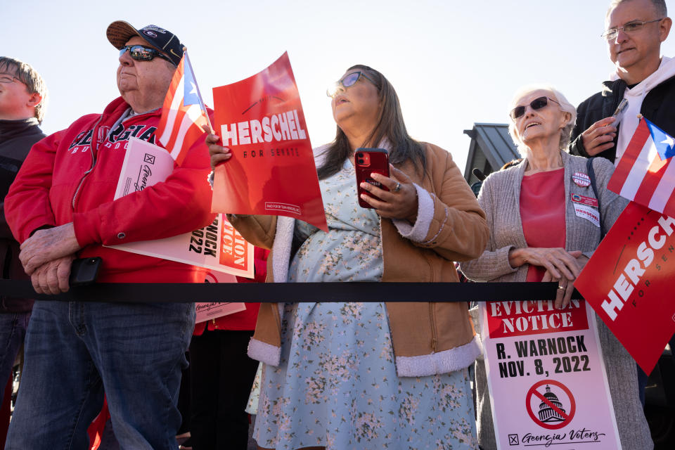 A half dozen people are visible behind a rope holding signs and handheld Puerto Rican flags. One sign reads Herschel for Senate. Another reads Eviction notice, R. Warnock, Nov. 8, 2022.