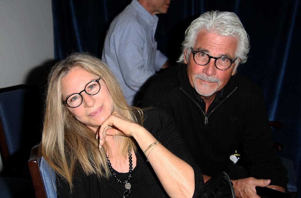 Barbra Streisand, in a black shirt and glasses, smiles at the camera, while husband Jame Brolin, also in a black shirt and glasses, sits behind her.