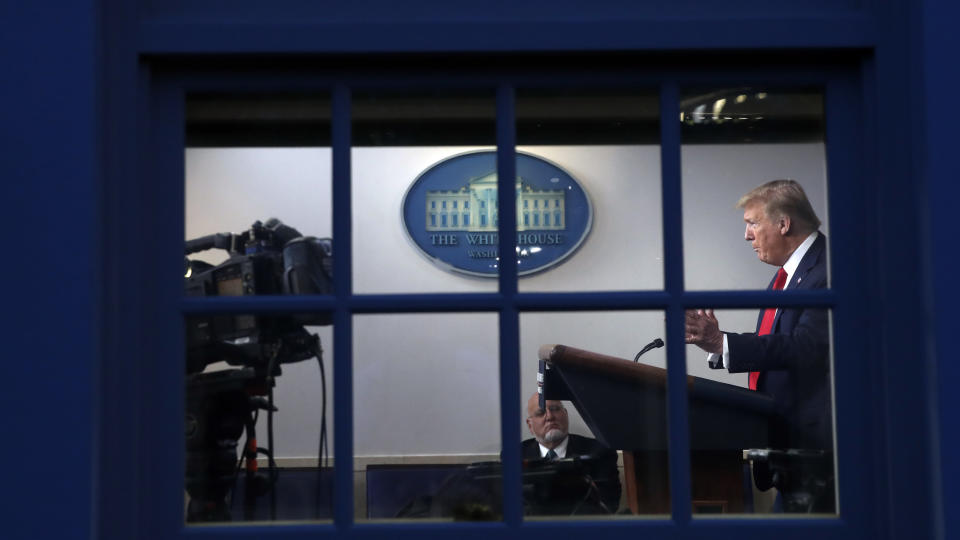 As seen through a window, President Donald Trump speaks about the coronavirus in the James Brady Press Briefing Room of the White House, Friday, April 17, 2020, in Washington. Public health experts are becoming concerned that people will use their partisan identification to decide which officials to believe whenever parts of the country start edging back to a new normal. (AP Photo/Alex Brandon)