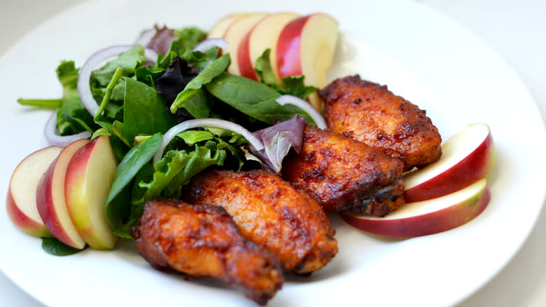 Four glazed apple cider wings on a plate surrounded by a bed of greens and apple slices