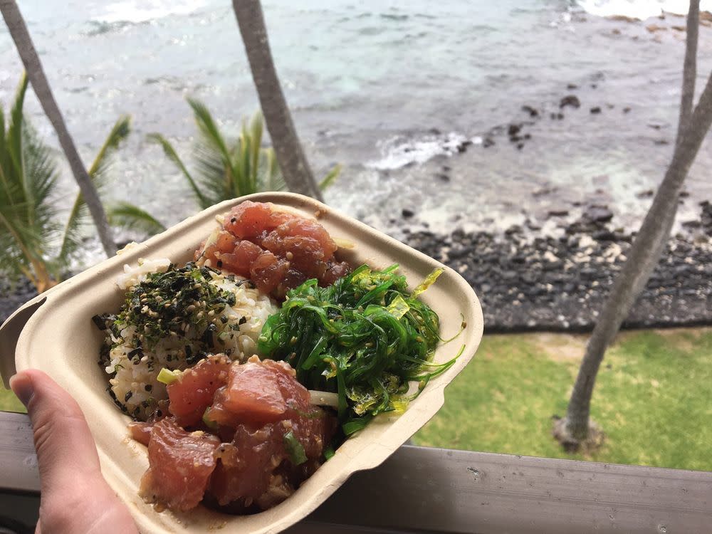 Person's hand holding bowl of poke in front of beach in Hawaii