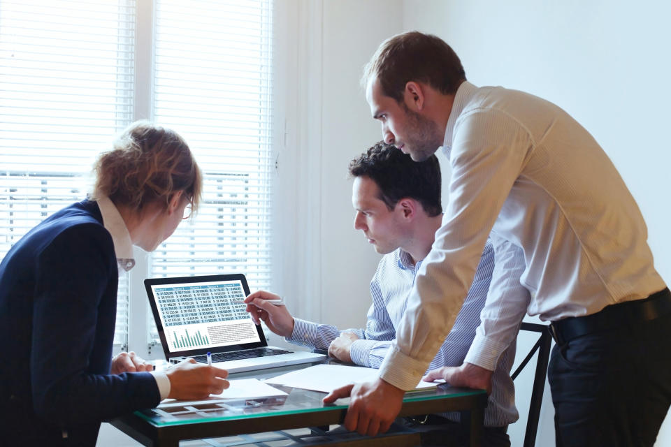 A group of three office workers are gathered around a computer, looking at a chart.