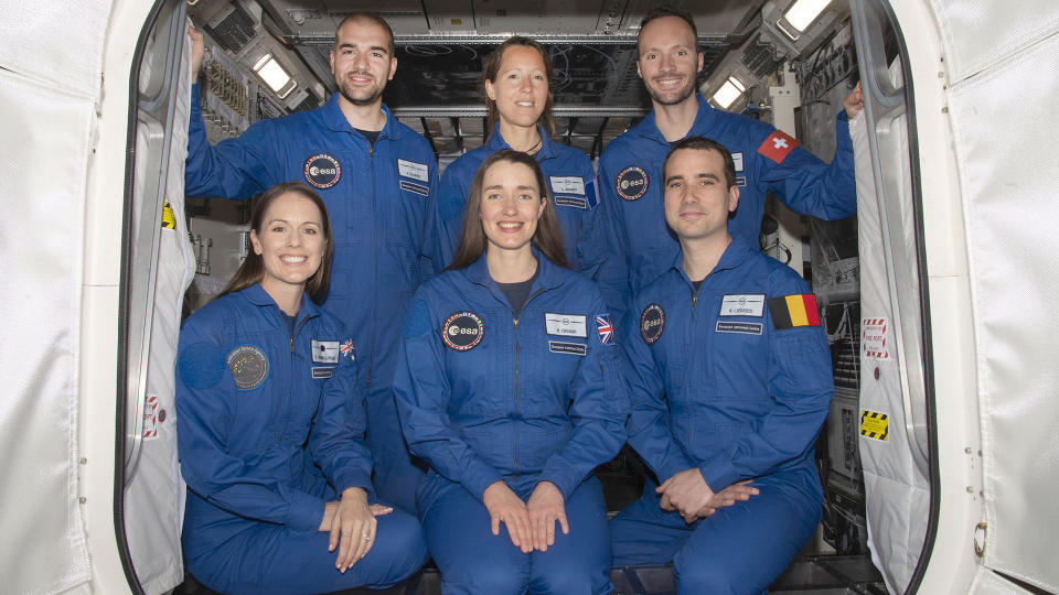 six people in blue flight suits pose in the doorway of a simulated space module