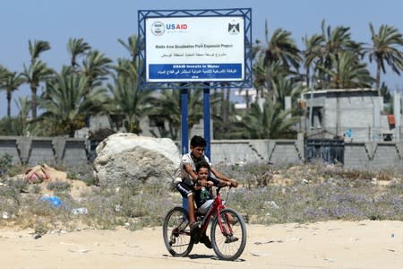 Palestinian boys ride a bicycle past a sign for USAID project for Gaza desalination plant, in the central Gaza Strip