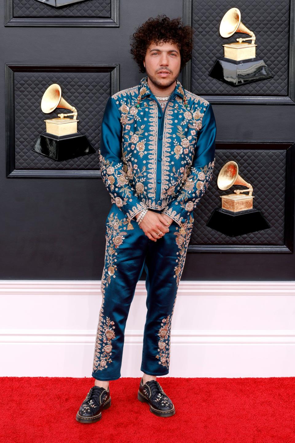 Benny in a turquoise blue satin jacket and pants with gold floral embellishments throughout. He's wearing ankle Doc Marten shoes with floral designs on it.