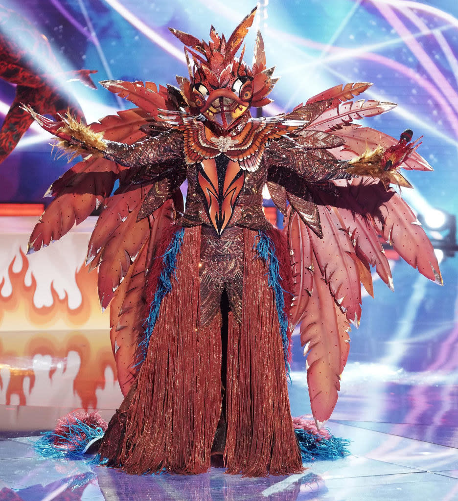 still from Season 5 of "The Masked Singer" featuring a guest in a bizarre phoenix-like costume complete with feathers and lengthy fringe