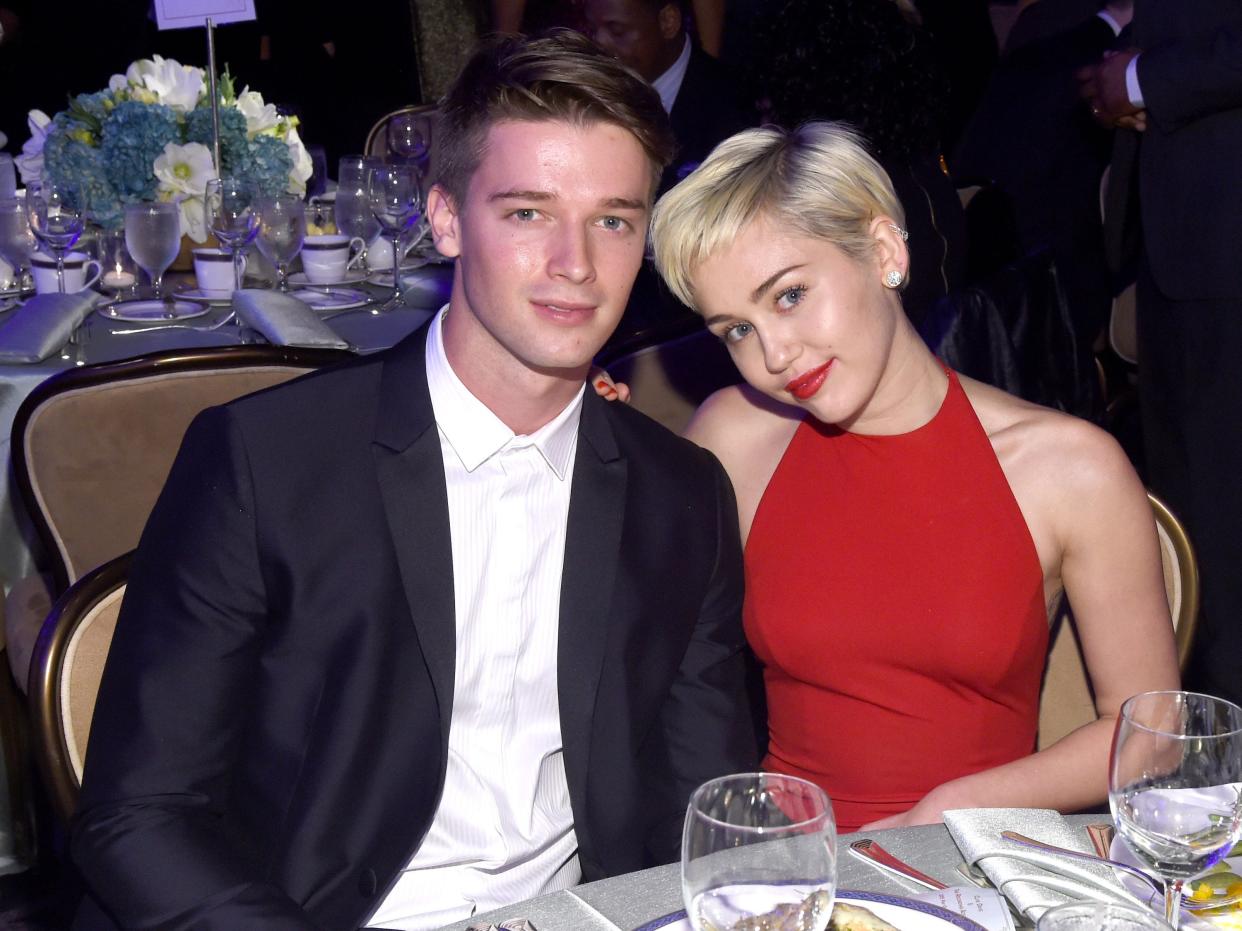 Patrick Schwarzenegger, in a black suit and white shirt, poses for photos with Miley Cyrus, who has short blonde hair and is wearing a red dress with red lipstick, at a 2015 event.