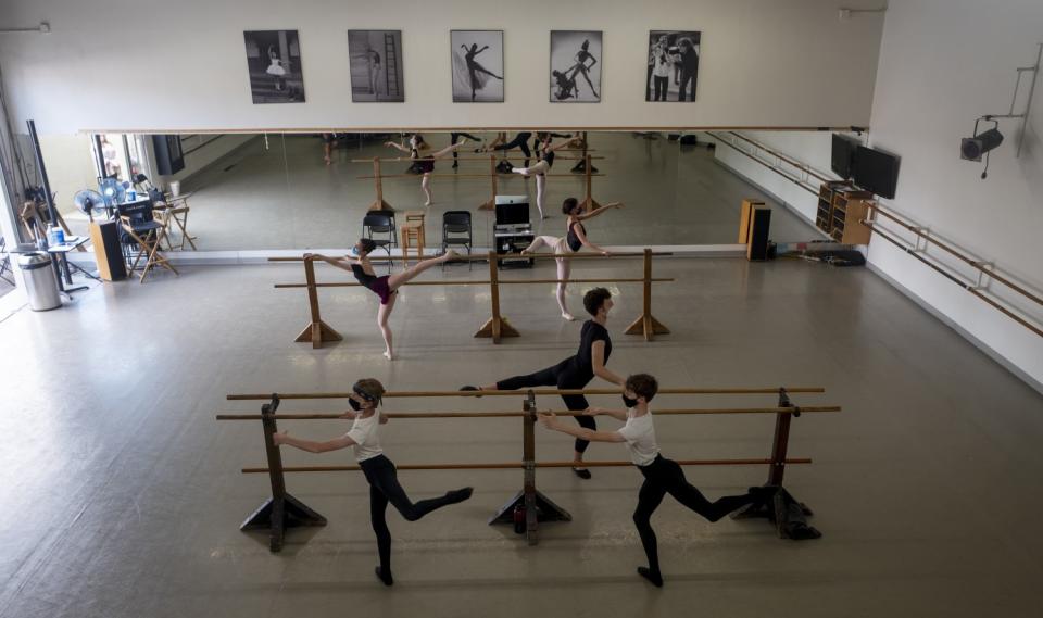 Five young dancers wearing masks rehearse at a barre in a dance class.