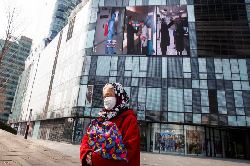 A woman wearing a face mask walks past a screen showing a video about protective measures against the new coronavirus in Beijing