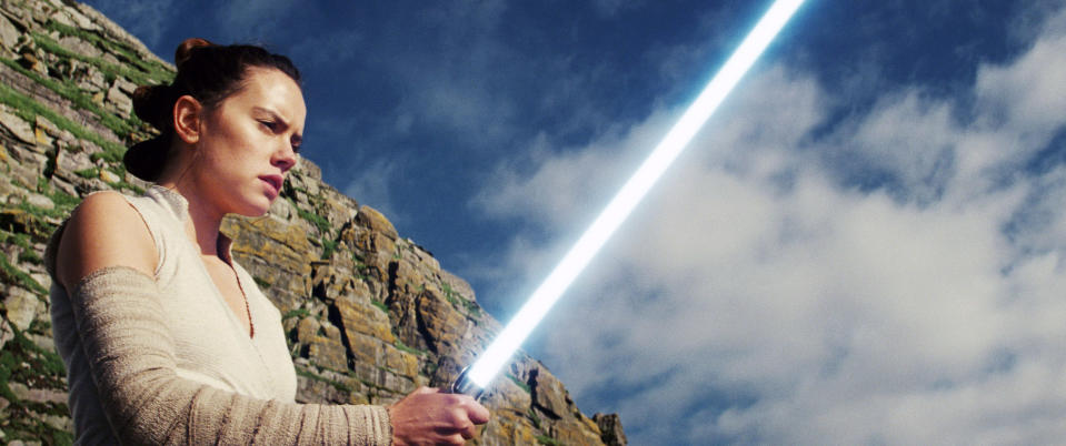 Rey from Star Wars appears to be concentrating as she holds her lightsaber on top of a rocky area.