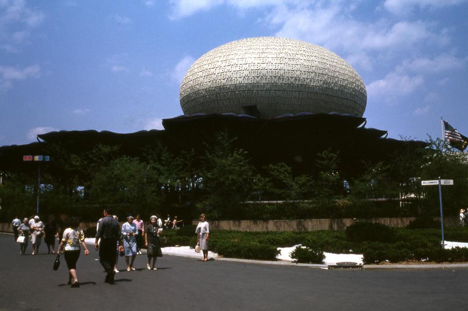 A white ovoid pavilion covered in textured letters rises above a pedestrian path at the New York World's Fair in 1964.