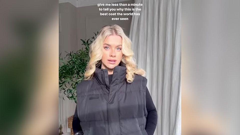 A still from one of the ads banned by the ASA. Grace, a 27-year-old woman with blonde hair, wears a black coat, her hands in the pockets. Text above her reads: "Give me less than a minute to tell you why this is the best coat the world has ever seen".