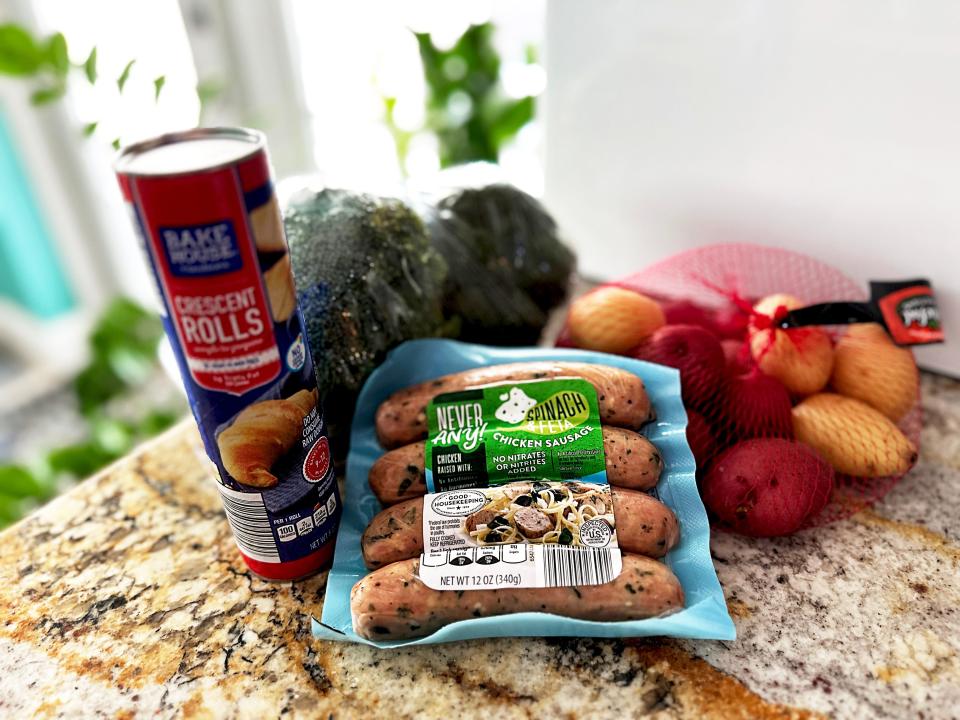 A pack of sausages with blu packaging, a bag of potatoes, broccoli, and a red and blue can of crescent rolls on a stone countertop