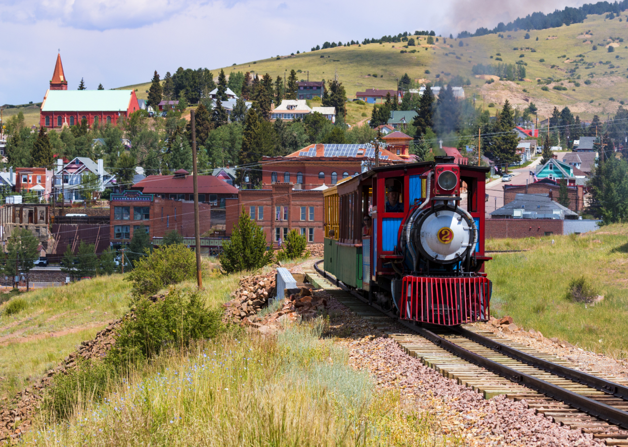 Train in motion on railroad in the foreground with the town of Cripple Creek, Colorado in the background, with mountains, on a sunny day