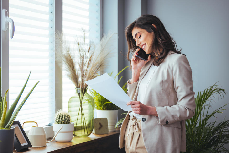 Beautiful business woman wearing modern suit, talking smartphone and holding papers in hands. Open space loft office. Windows background. Horizontal mockup.