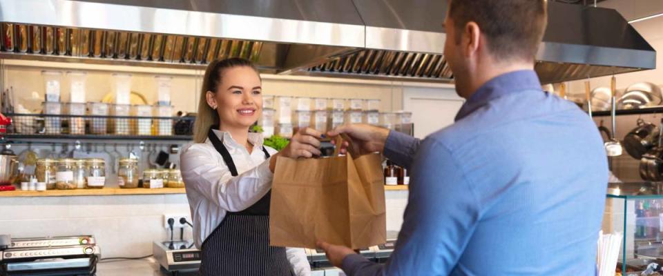 Cheerful waitress wearing apron serving customer at counter in restaurant - Small business and service concept with young business owner woman giving bag with takeaway food to client