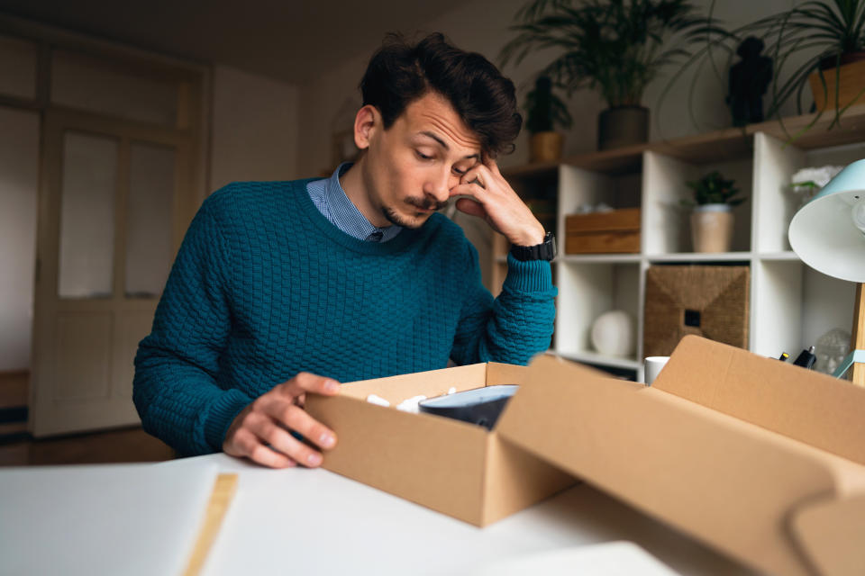 A young Caucasian man sits at home and is disappointed after unpacking a parcel with the wrong order. Online shopping concept.