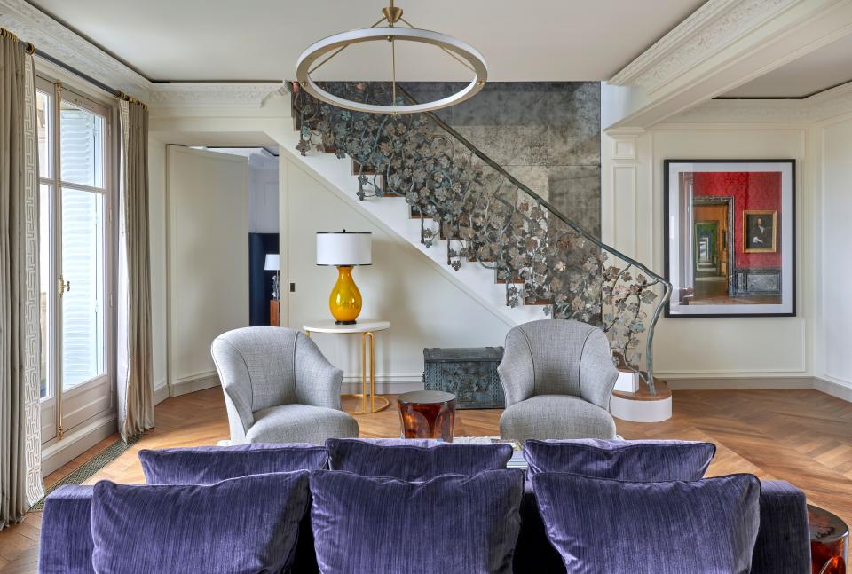 Another view of the main salon, which opens into the main bedroom through the door at left. The custom purple double sofa is in Dedar velvet, and the shell chairs are by Doghia in Rubelli fabric. The yellow mercury glass lamp under the steps is Pinto Paris. Wood notes, “The Halo chandelier was used to keep a freshness to the space and remind us that is is a modern apartment.” The ornate stair railing leads up to the library, and the photograph is by artist Robert Polidori from his Versailles series.