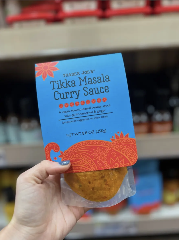 Hand holding a jar of Trader Joe's Tikka Masala Curry Sauce in front of grocery shelves