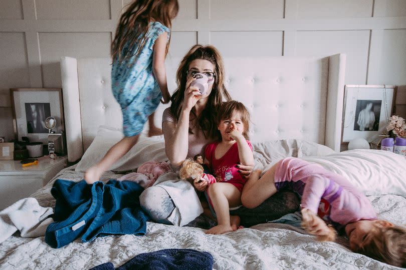 Photo of stressed out young mother sipping on a cup of coffee on her messy bed while her three daughters are playing around her.