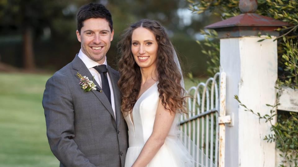 2021 MAFS couple Belinda and Patrick stand near a garden gate on their show wedding day. He wears a grey suit and she wears a gown with a plunging neckline with her long curly hair flowing free. Photo: Nine Network