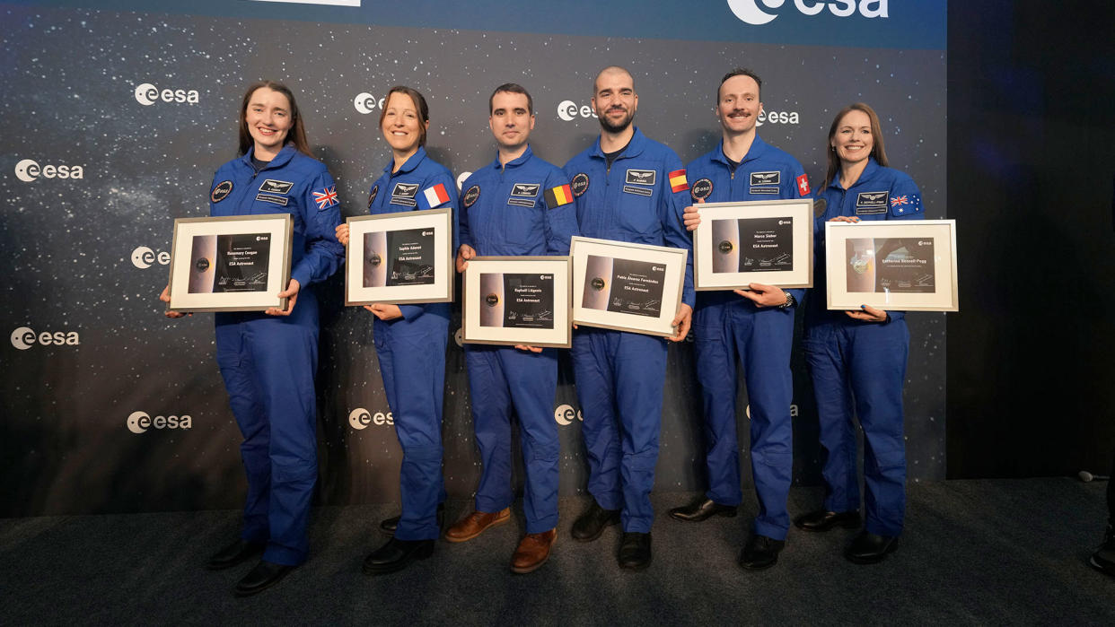  Six people in blue flight suits stand on a stage, each one holding a plaque. 