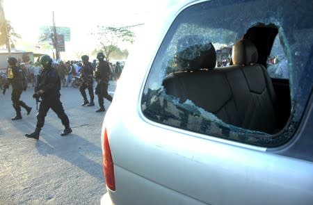 Riot police officers walk past a damaged car during a protest in Makassar