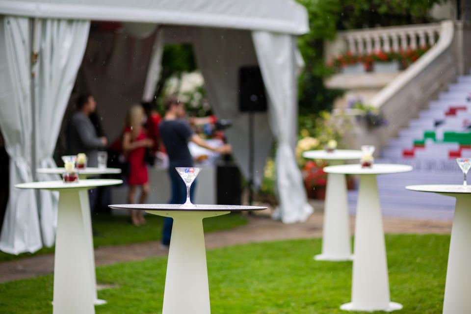Outdoor wedding reception with high tables set for cocktails. Guests in background include a woman in a red dress and a man in a blue shirt