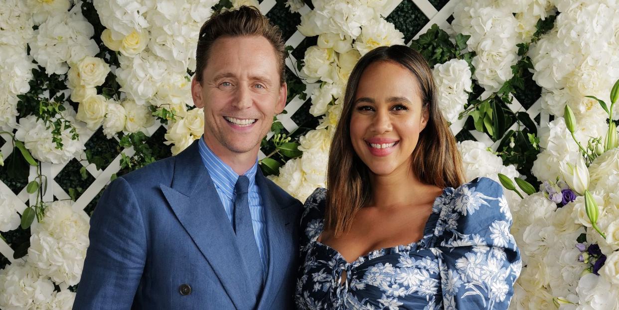 tom hiddleston wearing a blue suit and tie, standing in front of a trellis of white hydrangea flowers next to zawe ashton, who's wearing a long blue and white floral dress, at wimbledon in july 2023